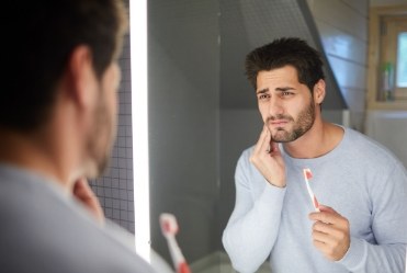 Man with toothbrush looking in mirror while holding cheek in pain